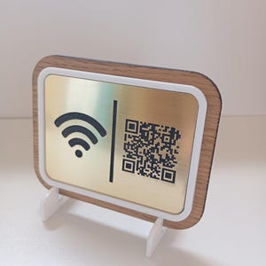 Può includere: Un cartello rettangolare con una piastra metallica dorata che presenta un simbolo Wi-Fi nero e un codice QR, incorniciato da un bordo bianco e un profilo effetto legno. Il cartello è sostenuto da due piccoli supporti bianchi dal design moderno.