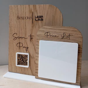 Może przedstawiać: Drewniany szyld z dwoma łukowatymi panelami z napisem Bespoke Laser Lab, Scan to Pay oraz Price List. Lewy panel zawiera kod QR w białej ramce, a prawy panel posiada puste białe kwadratowe pole. Całość umieszczona jest na białej podstawie.