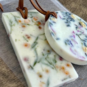 May include: Two white, rectangular and circular, scented wax pendants with embedded dried flowers and herbs. The pendants are attached to brown leather cords. The rectangular pendant is approximately 2 inches long.