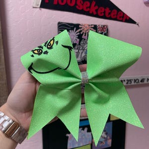 May include: A green glitter cheer bow with a black and white Grinch face design.