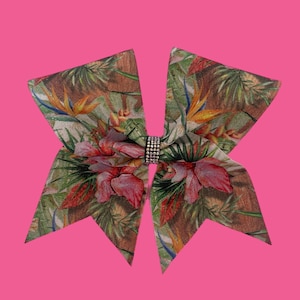 May include: A large, decorative hair bow featuring a tropical floral pattern with pink, orange, and green hues. The bow is accented with a sparkling, silver-colored rhinestone band at the center. The bow is set against a bright pink background.
