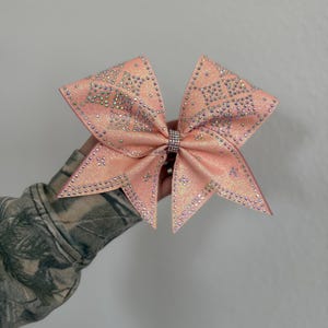 May include: A large, peach-colored hair bow with a glittery finish, decorated with numerous clear, reflective rhinestones arranged in a geometric pattern across the fabric. The center of the bow is cinched with a band of small, sparkling crystals.