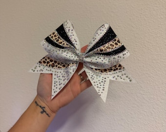Pode incluir: Um laço de cabelo grande com um design em camadas, apresentando seções de tecido branco, preto e com estampa de leopardo. Toda a superfície é densamente adornada com strass transparentes brilhantes e o centro é preso por uma faixa de cristais prateados.