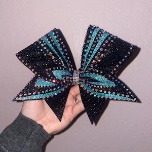 May include: Black glitter cheerleading bow with turquoise accents and iridescent rhinestones. The bow features a central rhinestone embellishment and is held in a hand against a pink background.