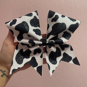 May include: A large, decorative hair bow with a cow print design. The bow features a white background with black spots and a black center. The bow is made of a glittery material.