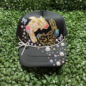 May include: Black trucker hat with a koi fish design. The hat features a white and gold koi fish with pink accents, surrounded by pearls and iridescent sequins. A white string of pearls and a bow adorn the brim.