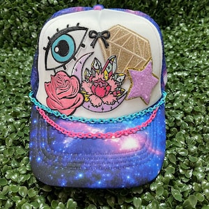 May include: A white and blue trucker hat with a galaxy print. The hat has a variety of colorful patches including a pink rose, a blue eye, a pink star, a gold diamond, and a pink and purple flower. The hat also has a pink and blue chain around the brim.