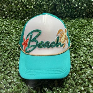 May include: A teal and white trucker hat with the word "Beach" in green glitter, a gold turtle, and a red starfish. The hat has a teal brim and a white front panel, with a gold rope detail.