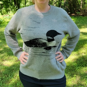 May include: A light gray long-sleeve crewneck sweater featuring a large, detailed black and white graphic of a loon swimming on water. The knit garment has ribbed cuffs, a ribbed hem, and a ribbed neckline, providing a consistent texture throughout.