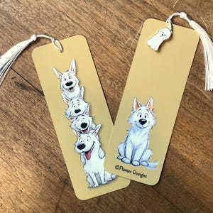 Può includere: Due segnalibri rettangolari con nappe bianche. Uno presenta una pila di cani bianchi dei cartoni animati, l'altro un singolo cane. I segnalibri sono beige con il testo "Flamax Designs".