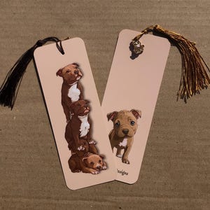 Può includere: Due segnalibri con illustrazioni di cuccioli di pitbull marroni e bianchi. Un segnalibro presenta tre cuccioli impilati, l'altro un singolo cucciolo. Ogni segnalibro ha un nappa, uno nero e uno dorato.