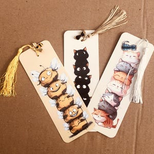 May include: Three whimsical bookmarks with tassel accents. One features a stack of smiling bees, another a tower of black cats, and the last a pile of colorful, sleeping kittens. Each bookmark is rectangular with a rounded top.
