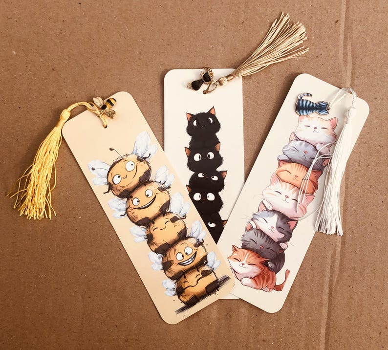 May include: Three whimsical bookmarks with tassel accents. One features a stack of smiling bees, another a tower of black cats, and the last a pile of colorful, sleeping kittens. Each bookmark is rectangular with a rounded top.