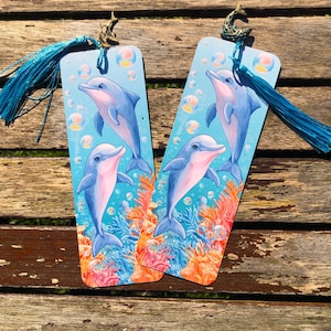 Könnte beinhalten: Zwei Lesezeichen mit Delfin-Motiv und blauen Quasten. Jedes Lesezeichen zeigt zwei Delfine in einer türkisfarbenen Ozeanszene mit Blasen und Korallen. Die Delfine sind blau und rosa, und die Quasten sind passend blau.