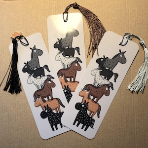 Könnte beinhalten: Drei weiße, rechteckige Lesezeichen mit einem Cartoon-Pferdedesign. Jedes Lesezeichen zeigt einen Stapel von Pferden in verschiedenen Farben, darunter Schwarz, Braun, Grau und Weiß. Die Lesezeichen haben Quasten in Schwarz, Braun und Silber.