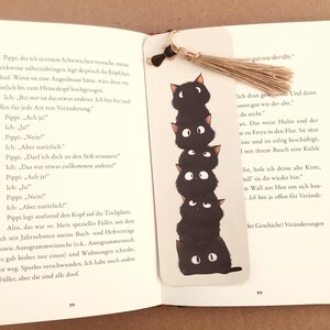 May include: A whimsical bookmark featuring a stack of six black cat illustrations with white eyes. The bookmark is made of white cardstock and has a natural twine tassel. The bookmark is placed in an open book.