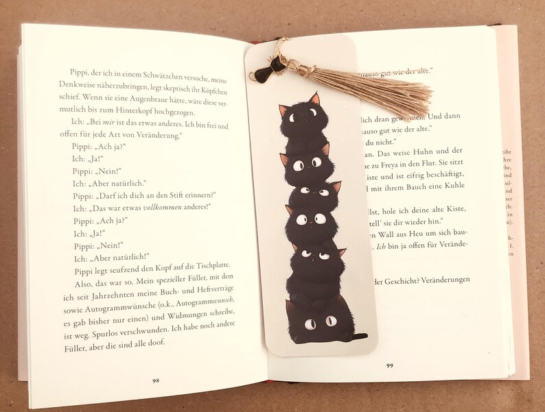 May include: A whimsical bookmark featuring a stack of six black cat illustrations with white eyes. The bookmark is made of white cardstock and has a natural twine tassel. The bookmark is placed in an open book.