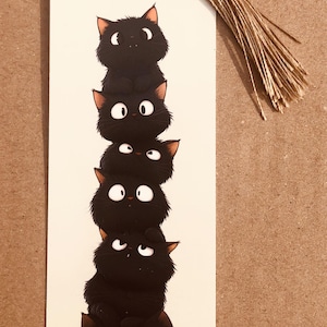 May include: A whimsical bookmark featuring a stack of six black cartoon cats with white eyes and orange ears. The bookmark has a tassel and a small black cat charm at the top.