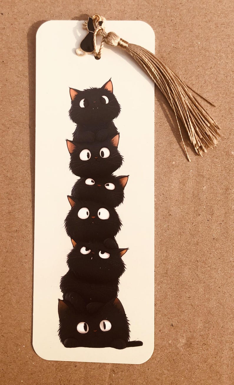 May include: A whimsical bookmark featuring a stack of six black cartoon cats with white eyes and orange ears. The bookmark has a tassel and a small black cat charm at the top.