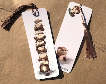 Könnte beinhalten: Zwei weiße Lesezeichen mit braunen Quasten. Ein Lesezeichen zeigt einen Stapel von Ottern, das andere einen Otter im Wasser und einen kleinen Otter auf einem Surfbrett. Die Lesezeichen sind zum Lesen.