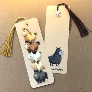 Könnte beinhalten: Zwei Lesezeichen mit Pferdemotiv. Ein Lesezeichen zeigt einen Stapel von fünf Cartoon-Pferden in verschiedenen Farben, während das andere ein einzelnes schwarzes Pferd mit weißen Punkten zeigt. Beide haben Quasten.