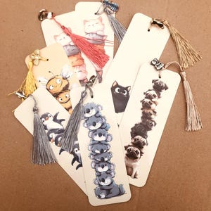 May include: A collection of decorative bookmarks featuring various animal illustrations. Designs include stacked cats, koalas, pugs, a penguin, a bee, and a black cat. Each bookmark has a tassel and a small charm at the top.