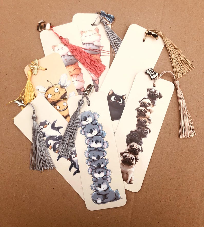 May include: A collection of decorative bookmarks featuring various animal illustrations. Designs include stacked cats, koalas, pugs, a penguin, a bee, and a black cat. Each bookmark has a tassel and a small charm at the top.
