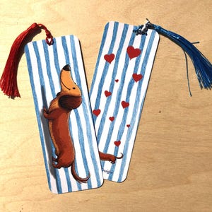 Può includere: Due segnalibri rettangolari in carta con un motivo a righe verticali blu e bianche. Uno mostra l'illustrazione di un bassotto marrone con una nappa rossa, l'altro presenta cuori rossi e una nappa blu attaccata nella parte superiore.