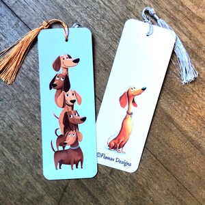 Può includere: Due segnalibri illustrati con cani bassotti. Un segnalibro ha una pila di bassotti, l'altro ha un singolo cane. Entrambi hanno nappe. Il testo "Flamax Designs" è su un segnalibro.