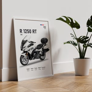 May include: A framed poster featuring a white BMW R 1250 RT motorcycle. The poster includes specifications and the BMW logo. A potted plant with large green leaves sits to the right of the poster on a wooden floor.