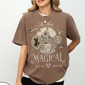 May include: Brown t-shirt with a vintage-style graphic featuring a castle, teacup, Mickey Mouse silhouette, and the words "The Most Magical Place on Earth." The design includes a globe and fireworks. The shirt has short sleeves.