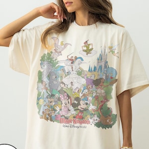 May include: Ivory-colored t-shirt featuring a colorful graphic of various Disney characters, including Mickey Mouse, Minnie Mouse, and Cinderella's castle. The text "Magic Kingdom Walt Disney World" is printed below the graphic.