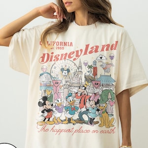 May include: Ivory-coloured t-shirt with a Disneyland graphic. The design showcases the Disneyland logo, park imagery, and Disney characters. Text includes "California Disneyland est. 1955" and "The happiest place on earth."