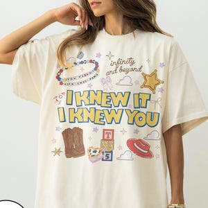 May include: An ivory t-shirt featuring a graphic print with the text "I KNEW IT I KNEW YOU" and "infinity and beyond". The design includes illustrations of cowboy boots, a red hat, alphabet blocks, a star, clouds, and a beaded necklace.