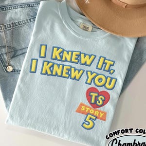 Puede incluir: Una camiseta de manga corta de color azul claro con texto bordado en amarillo que dice I KNEW IT, I KNEW YOU. Debajo del texto hay un coraz&oacute;n rojo con las letras TS, con la palabra STORY y el n&uacute;mero 5 impresos en amarillo debajo.