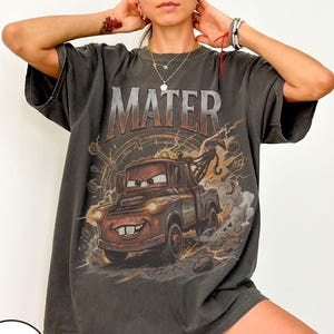 May include: Dark gray t-shirt featuring a graphic of Mater from the movie Cars. The word "MATER" is printed above the character. The design includes details like a tow hook and lightning bolts. The shirt has short sleeves and a relaxed fit.