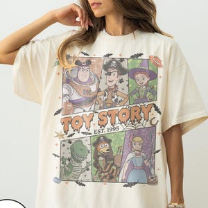 May include: An ivory short-sleeve t-shirt featuring a graphic print of Toy Story characters in Halloween costumes. The design includes Buzz Lightyear, Woody, Jessie, Rex, Slinky Dog, and Bo Peep, with the text "TOY STORY EST. 1995" and bats.