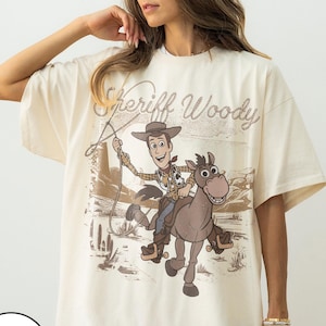 May include: Ivory-colored t-shirt featuring a graphic of Sheriff Woody riding a horse, with the text "Sheriff Woody" above the image. The design is in shades of brown and tan, with a desert landscape backdrop.