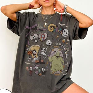 May include: Dark gray t-shirt featuring a colorful graphic design with characters and elements from "The Nightmare Before Christmas." The design includes Jack Skellington, Sally, Oogie Boogie, and other iconic imagery. The shirt has short sleeves and a relaxed fit.
