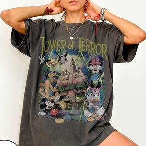 K&ouml;nnte beinhalten: Dunkelgraues T-Shirt mit einer Grafik von Disney-Figuren vor dem Hollywood Tower Hotel, mit dem Text "Tower of Terror" in Gr&uuml;n. Das Design umfasst Micky Maus, Minnie Maus, Donald Duck und Goofy. Das Shirt hat kurze &Auml;rmel.