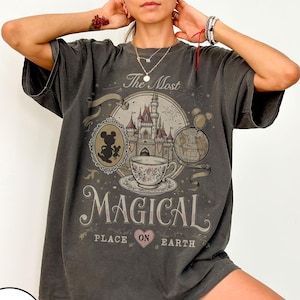 May include: Dark gray t-shirt with a graphic design featuring a castle, teacup, and Mickey Mouse silhouette. The text reads "The Most Magical Place on Earth." The shirt has short sleeves and a relaxed fit.
