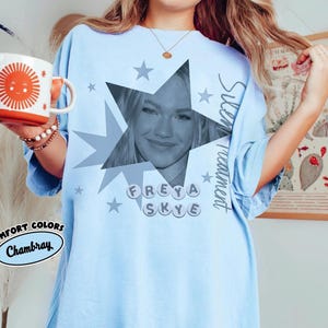 May include: Light blue t-shirt with a star graphic featuring a portrait and the name "FREYA SKYE". The shirt also has the text "Silent Treatment" and "Comfort Colors Chambray". A white and orange mug with a sun design is held in the image.