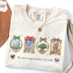 K&ouml;nnte beinhalten: Elfenbeinfarbenes T-Shirt mit vier gerahmten Illustrationen von Themenpark-Wahrzeichen, die jeweils mit einer Schleife verziert sind. Das Shirt enth&auml;lt den Text "The most magical place on Earth" und ist mit einer goldenen Halskette versehen.