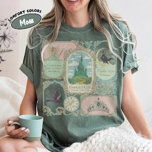 以下が含まれることがあります： モスグリーンのTシャツ。カラフルなグラフィックには、妖精、お城、ほうきに乗った魔女、帽子、時計、王冠が描かれています。Tシャツのテキストには「Emerald City」と「Together We're Unlimited」が含まれています。