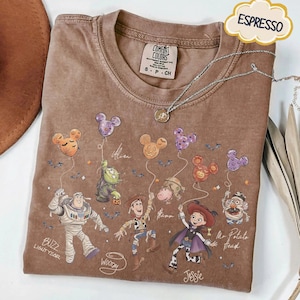 Pode incluir: Camiseta cor de caf&eacute; com personagens de Toy Story da Disney segurando bal&otilde;es em forma de Mickey Mouse. A camisa inclui Buzz Lightyear, Woody, Jessie e Sr. Cabe&ccedil;a de Batata. Um colar de prata com um pingente repousa sobre a camisa.