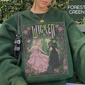 May include: Forest green sweatshirt with a graphic from the musical "Wicked." The graphic features two characters, one in a pink dress and the other in a black dress, with the word "Wicked" in green. The sweatshirt is a solid forest green colour.