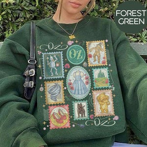Könnte beinhalten: Forstgrünes Sweatshirt mit einer Collage von Illustrationen im Briefmarkenstil aus Der Zauberer von Oz. Das Design umfasst Bilder vom Blechmann, Dorothys Kleid, dem Löwen und den roten Schuhen der bösen Hexe. Das Wort "OZ" ist ebenfalls enthalten.