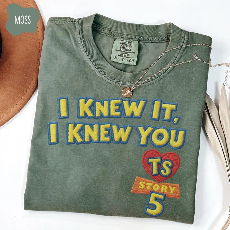 Puede incluir: Una camiseta de cuello redondo color verde musgo con texto bordado que dice I KNEW IT, I KNEW YOU, junto a un coraz&oacute;n rojo con las letras TS sobre la palabra STORY y el n&uacute;mero 5, todo en hilo de color amarillo y azul en el frente.