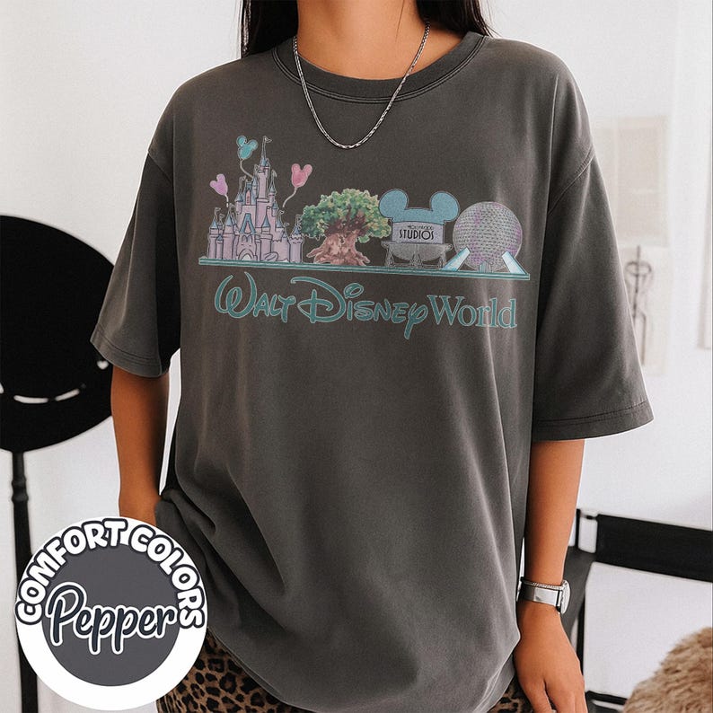 Puede incluir: Camiseta Comfort Colors gris oscuro con un dise&ntilde;o de Walt Disney World. El dise&ntilde;o incluye un castillo, un &aacute;rbol y las palabras "Walt Disney World" en turquesa. La camiseta tambi&eacute;n tiene las palabras "Comfort Colors Pepper" en un dise&ntilde;o circular.