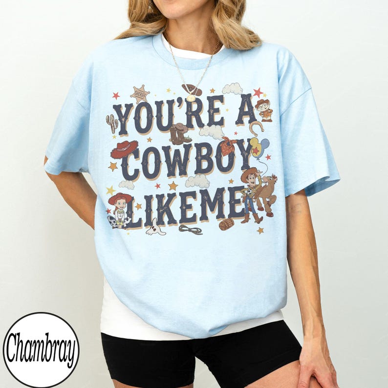 May include: A light blue short-sleeve t-shirt featuring the dark blue text "YOU'RE A COWBOY LIKEME" surrounded by various western-themed illustrations including a sheriff badge, cowboy hat, boots, clouds, stars, and cartoon characters.